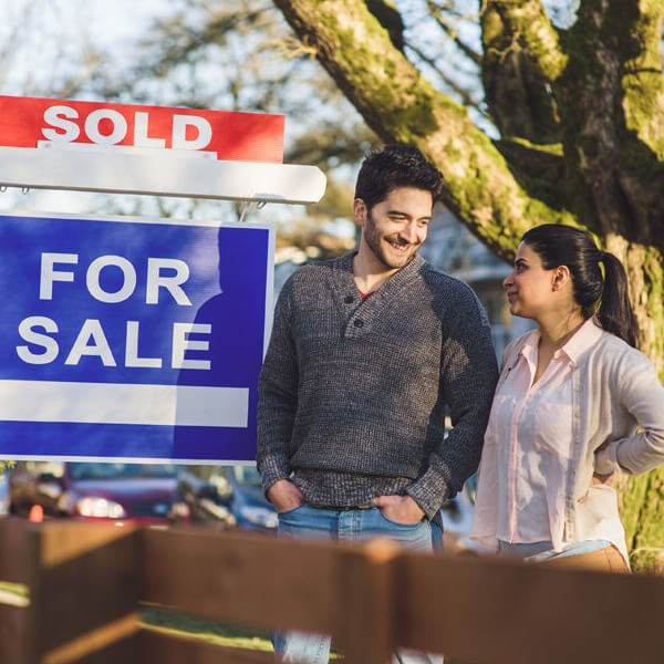 Couple smiling in front of house with a sign saying sold